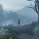 A young girl stands in front of flowers and a tree in a foggy landscape.